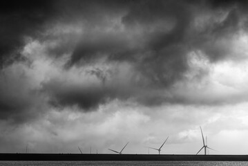 Dramatic black and white image of a dark stormy sky with thick clouds over a coastal landscape featu wind turbines on the horizon under ominous weather conditions