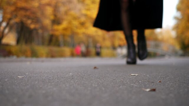 Confident autumn walk, dark boots with square heels pass close to lens, fallen leaves and soft light enrich seasonal mood.