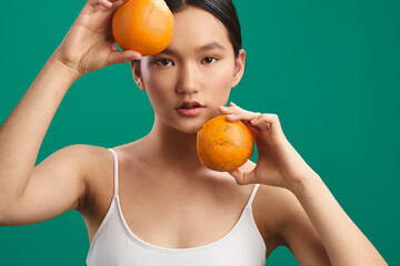 Asian woman holds oranges against a green backdrop, creating a fresh, vibrant portrait. She looks toward the camera with a calm, engaging expression.
