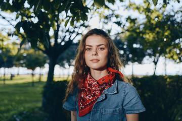 Fototapeta premium young woman in denim shirt with red bandana standing outdoors in sunny park surrounded by trees and green grass on summer day
