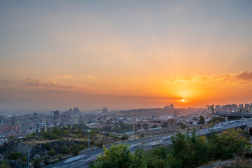 The sun setting over Yerevan, Armenia with radiant sunbeams and warm orange tones. The city skyline is bathed in soft light, creating a calm and picturesque urban sunset