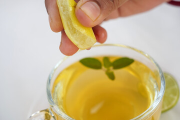 A woman hand squeeze lemon in the green tea. Ginger, half a lemon, honey and basil leaves in a bowl on a burlap top view. Useful vitamin ingredients in a cup of tea on a white background.