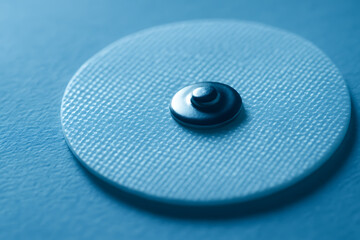 Close-up macro image of a medical electrode pad, showing detailed texture of the adhesive surface and the central metal connector. Captured in soft blue tones, it highlights the clean