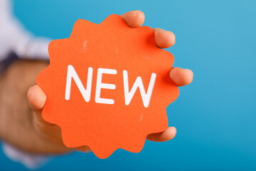 A hand holding a bright orange NEW label against a vibrant blue background, showcasing a fresh new product or concept. Ideal for marketing, prom