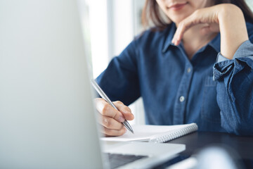 Asian woman remote working on laptop computer at home office. Business woman online meeting via laptop and writing on notebook on office table. Student studying online class, e-learning, close up