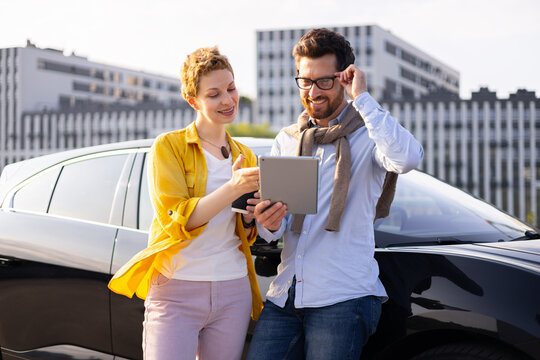 A man and woman stand near a car, looking at a tablet together, likely discussing something related to the vehicle or their journey.