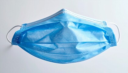 Blue disposable surgical face mask with pleated layers and white elastic ear loops, lying flat on a neutral surface, representing personal protective equipment and public health safety.