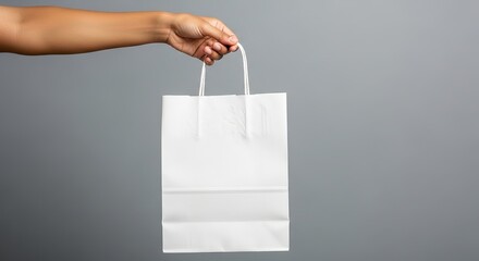 Arm holds a plain white paper shopping bag against a neutral gray backdrop in a studio setting.