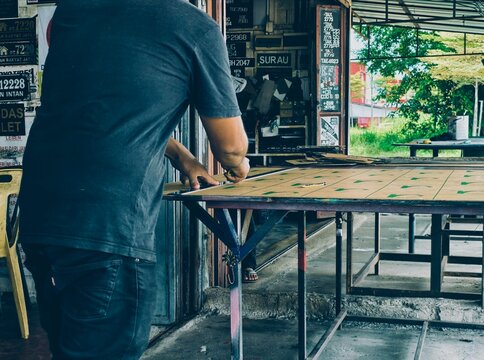 Manual worker precisely measuring and cutting a large sheet of material on a workbench at an outdoor workshop.
