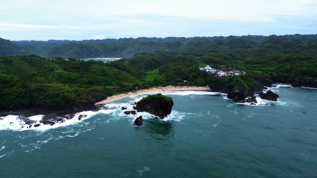 A stunning aerial view of a rocky beach surrounded by green hills and blue ocean waves crashing against the cliffs in Gunung Kidul on June 1, 2025.