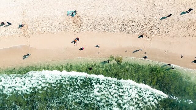 Aerial view of a tropical beach with gentle white waves lapping against golden sand and tourists relaxing by the sea in Gunung Kidul on June 1, 2025.