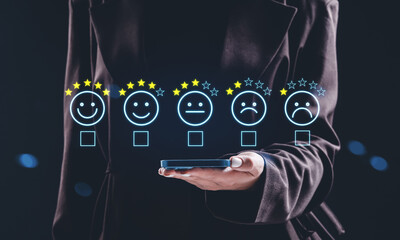 Businessperson holding smartphone projecting holographic customer satisfaction rating concept with five emoticons and stars on dark background.