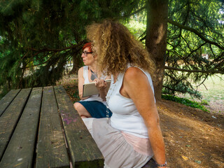 Two women enjoying a pleasant conversation while sitting at a rustic wooden picnic table in a sun-dappled park surrounded by lush green trees and foliage offering a serene outdoor ambiance for