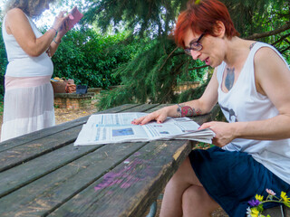 Two women in casual clothing enjoying a sunny day outdoors reading a newspaper and looking at a smartphone in a park setting