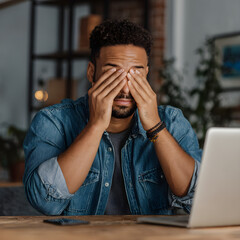 Young man covering his eyes with his hands while sitting at a desk with a laptop