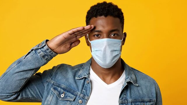 A young African American man wearing a denim jacket and a medical face mask salutes with a serious expression against a yellow background a perfect concept for frontline workers