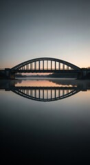 Naklejka premium An arched metal bridge with a silhouette of people walking across it, reflected in the still water below during a moody sunset