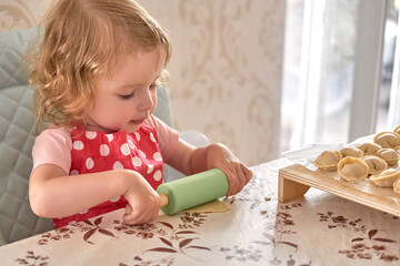 Little girl in red polka dot apron rolls dough with a green rolling pin.
Toddler learns cooking skills during fun home baking activity.
Cute child enjoys hands-on play and early culinary experience.
