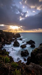 Coastal vista showcasing dramatic sky with sunburst over the ocean, rocky shore, and long exposure effect