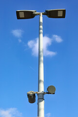 A tall street light pole with two large modern luminaires against a bright blue sky. Lower on the pole, two additional devices are mounted, possibly sensors or cameras.