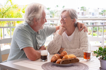 Smiling senior retired couple enjoying breakfast together sitting outside on home terrace with coffee, tea and baked goods. Peaceful retirement lifestyle.