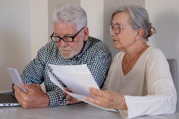 Senior couple sitting at home table with paper documents using laptop. Elderly man and woman reading documents, paying bills, managing bank finances, calculating taxes, debt, insurance, pension