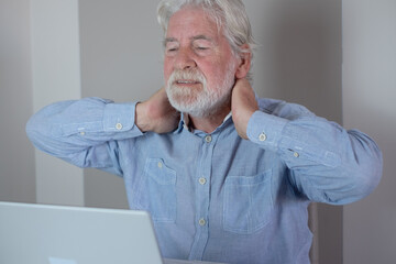 Senior man touching himself massaging his neck for painful bad posture while working on a laptop at...