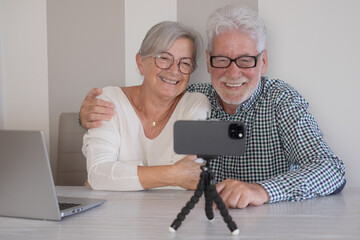 Video call concept. Elderly happy couple sitting at home talking remotely with family or friends on mobile phone webcam, modern senior  couple smiling sharing good news with distant people