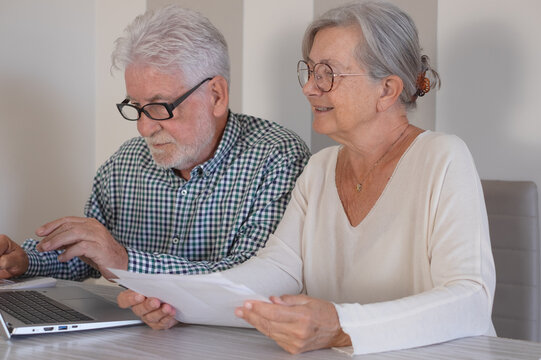 Senior couple sitting at home table with paper documents using laptop. Elderly man and woman reading documents, paying bills, managing bank finances, calculating taxes,