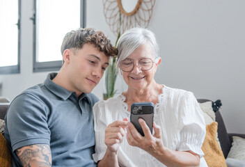 Happy grandmother learning how to use mobile phone technology from young grandson, handsome smiling boy teaching grandma shop online, book travel and vacation