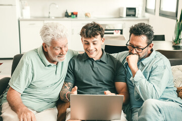 Three generations of men, grandfather, father, and son sitting together on sofa, laughing and enjoying time while using laptop at home, symbolizing family bonding and connection across generations