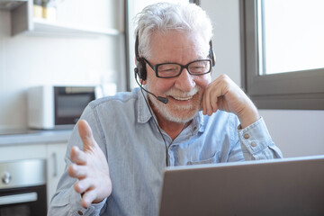 Video call concept. Senior caucasian bearded man wearing headphones smiling gesturing with hands while working online on laptop from home