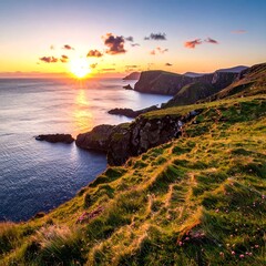 Fototapeta premium Coastal landscape at sunset, with vibrant orange and yellow hues from the sun. Rocky cliffs meet the vast blue ocean. Green grassy foreground