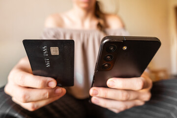 A young Caucasian woman holds a smartphone in one hand and a credit card in the other. She is engaged in online shopping or payment activity.