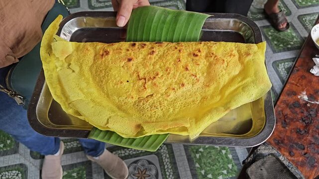 Vietnamese crispy pancake called Banh Xeo served on a metal tray lined with banana leaf, held by a person at a local eatery in Vietnam.