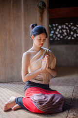 a young woman in traditional lanna attire sitting gracefully on a woven mat inside an old wooden temple in chiang mai, hands together in a wai gesture, eyes closed in calm meditation and serenity.