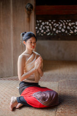 a young woman in traditional lanna attire sitting gracefully on a woven mat inside an old wooden temple in chiang mai, hands together in a wai gesture, eyes closed in calm meditation and serenity.