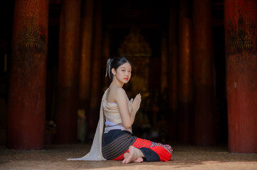 a young woman in traditional lanna costume sitting respectfully on the floor of a wooden temple in Chiang Mai, hands pressed together in wai gesture, surrounded by ancient red pillars and soft golden 