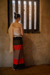 a young woman wearing traditional lanna dress standing quietly by a wooden window inside an old temple in chiang mai, warm sunlight casting soft shadows on the textured wall and woven bamboo floor.