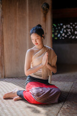 a young woman in traditional lanna attire sitting gracefully on a woven mat inside an old wooden temple in chiang mai, hands together in a wai gesture, eyes closed in calm meditation and serenity.