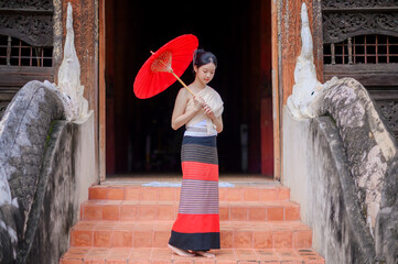 a young woman in traditional lanna northern thai costume holding a red umbrella stands gracefully on the temple stairs in Chiang Mai, surrounded by intricate wooden carvings and ancient naga sculpture
