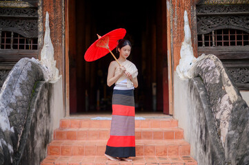 a young woman in traditional lanna northern thai costume holding a red umbrella stands gracefully on the temple stairs in Chiang Mai, surrounded by intricate wooden carvings and ancient naga sculpture