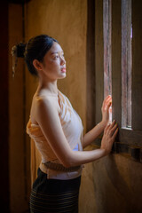 a young woman wearing traditional lanna dress standing quietly by a wooden window inside an old temple in chiang mai, warm sunlight casting soft shadows on the textured wall and woven bamboo floor.