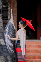 a young woman in traditional lanna northern thai costume holding a red umbrella stands gracefully on the temple stairs in Chiang Mai, surrounded by intricate wooden carvings and ancient naga sculpture