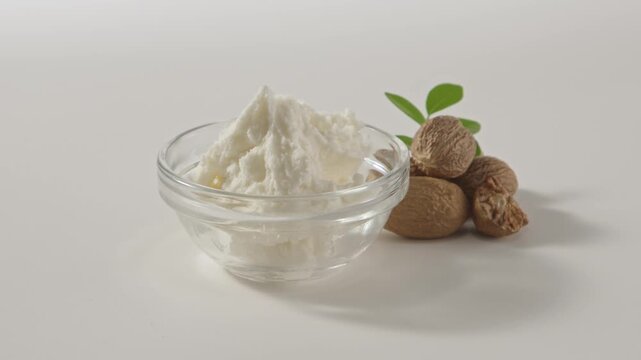 Glass bowl filled with creamy shea butter placed with raw nuts, styled for skincare cosmetic ingredient presentation in clean setup.