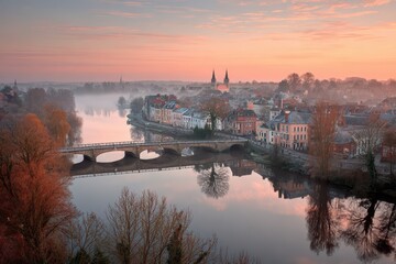 Misty European town, river reflects pastel sunrise sky and old bridge.