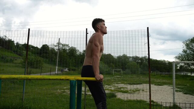 Strong man trains abdominal muscles raising legs on parallel bars at sports ground. Young shirtless sportsman exercising at nature. Sporty athlete working out at nature. Concept of sportive lifestyle