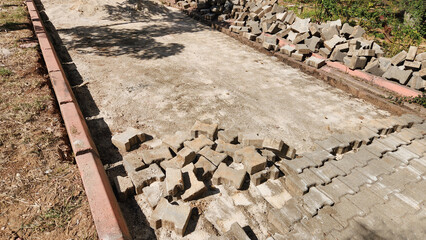 A pedestrian walkway construction site with a partially paved street bordered by bricks, featuring scattered interlocking pavers and a pile of additional stones
