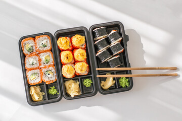 Assorted sushi rolls in black takeout boxes with chopsticks, wasabi and ginger on white background, showcasing fresh Japanese cuisine perfect for restaurant menus, delivery ads and food blogs