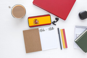 Flat lay with Spanish flag on smartphone, notebooks, coffee cup, and stationery on white desk, symbolizing language learning, online education, cultural communication between Spain English speakers © Evgeniia Primavera